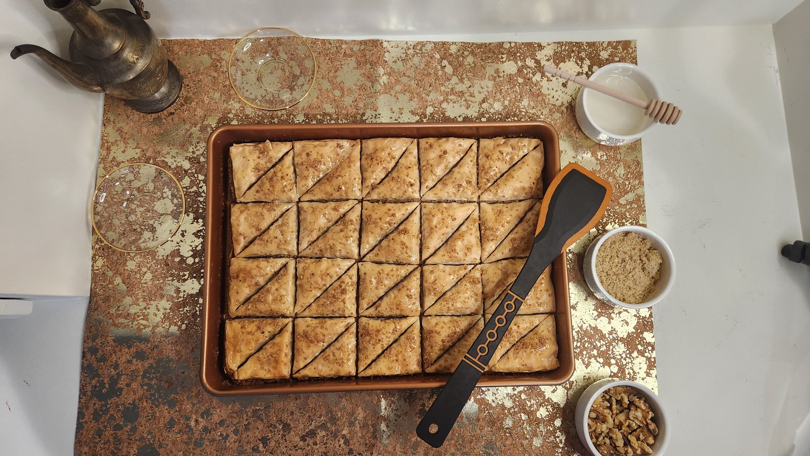 Overhead view of a full tray of honey-drizzled baklava with honey, walnuts, and tea on the side