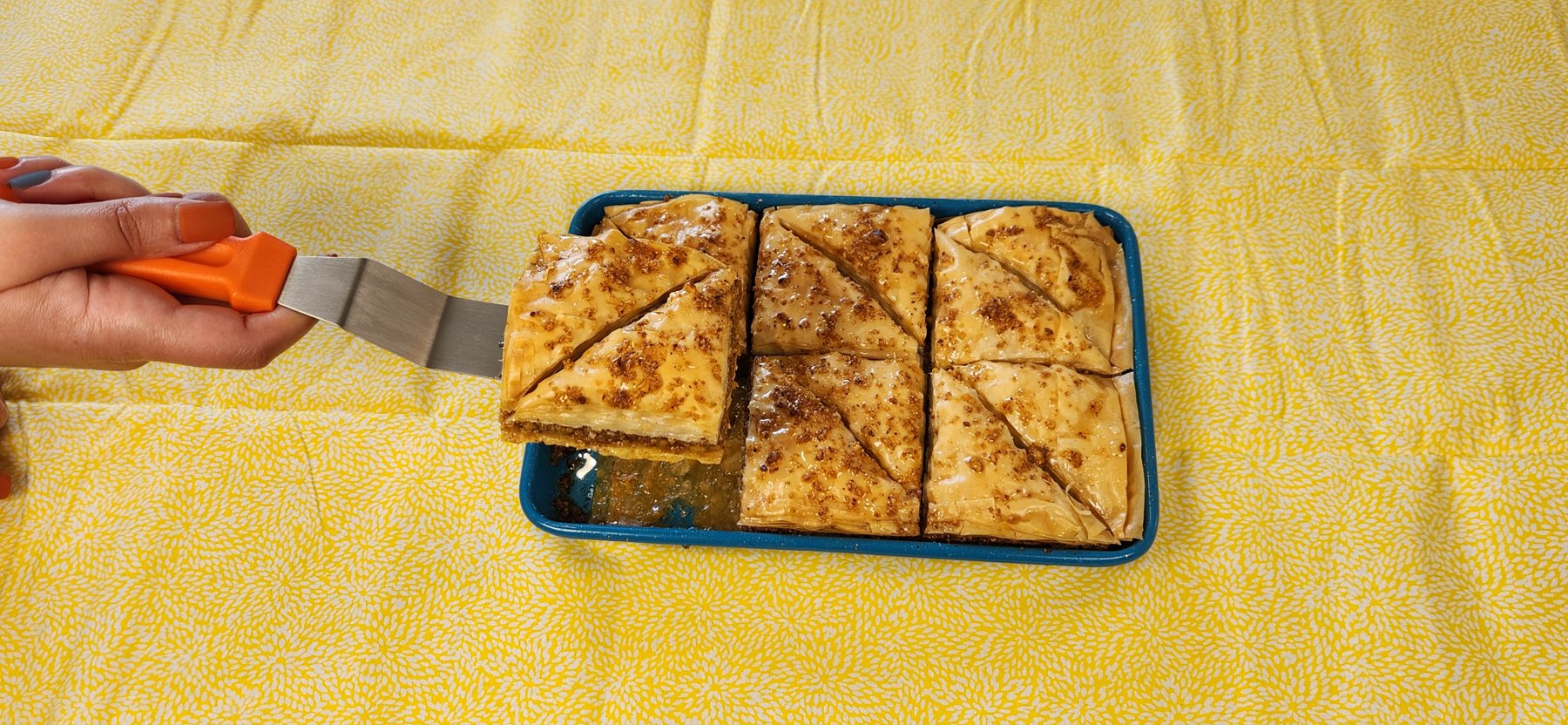 A hand lifting a glazed piece of baklava from a blue baking tray